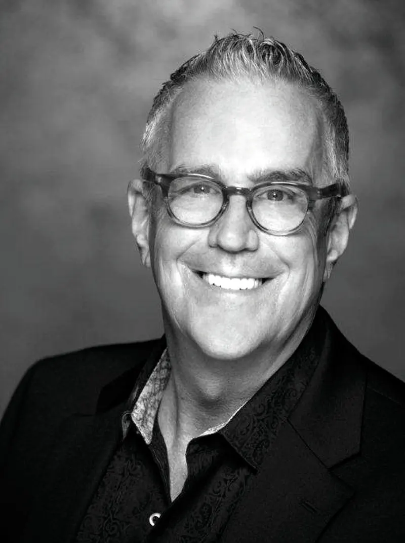 Smiling man with short hair and glasses, wearing a patterned shirt and blazer, ready to dazzle at the salon event. - Intercoiffure Canada America